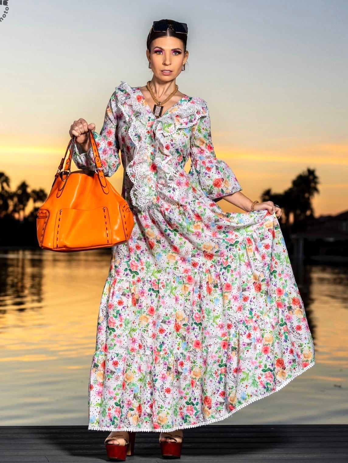 Woman in a couture long sleeve floral dress with front ruffle holding an orange bag by a waterfront at sunset