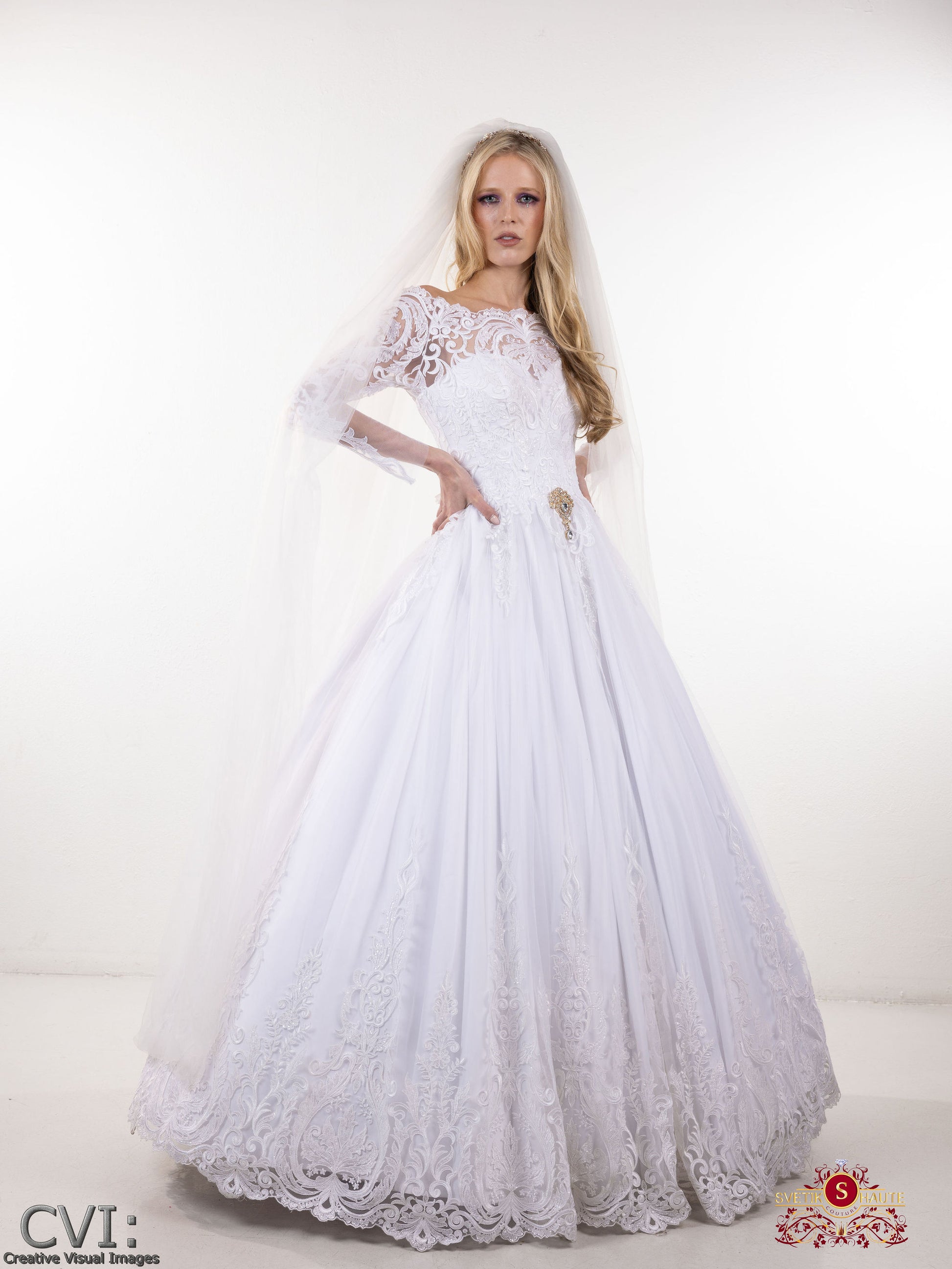 White lace couture wedding dress long sleeve and a veil on a white background on woman with long blonde hair and hands on her hips